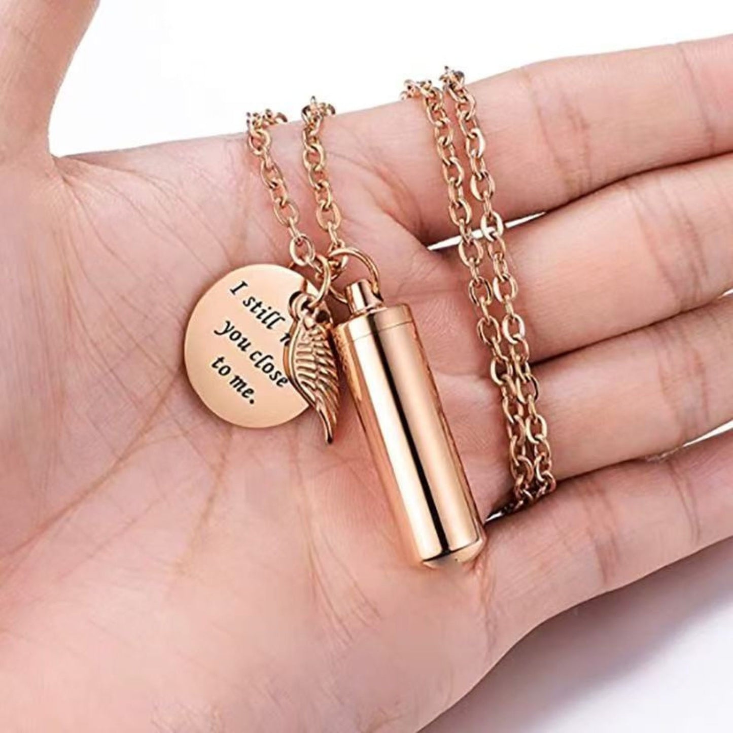 Hand holding rose gold pet urn necklace with wing charm and engraved round tag “I still need you close to me.”