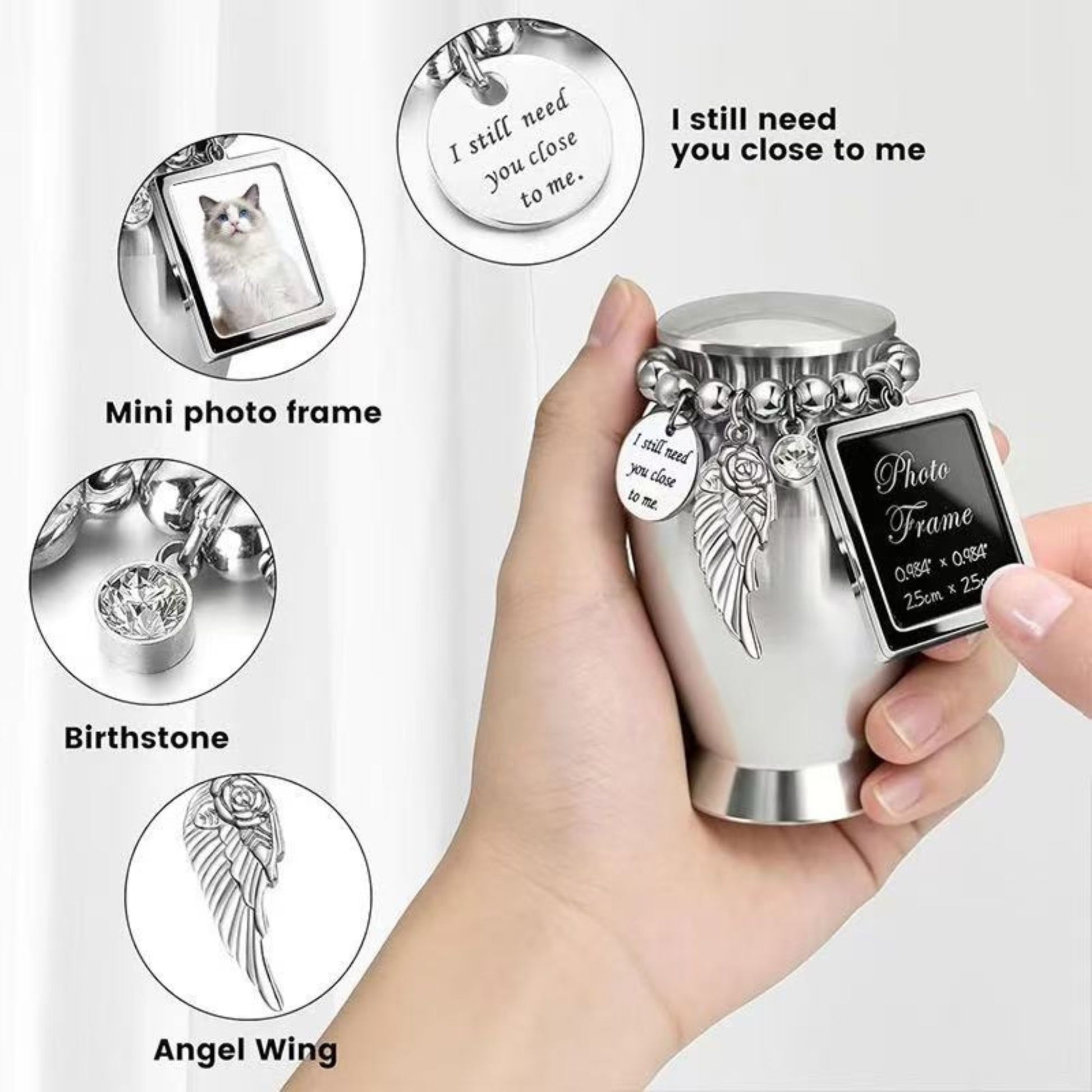 Hand holding small silver pet urn showing photo frame charm, angel wing charm, and “I still need you close to me” disc